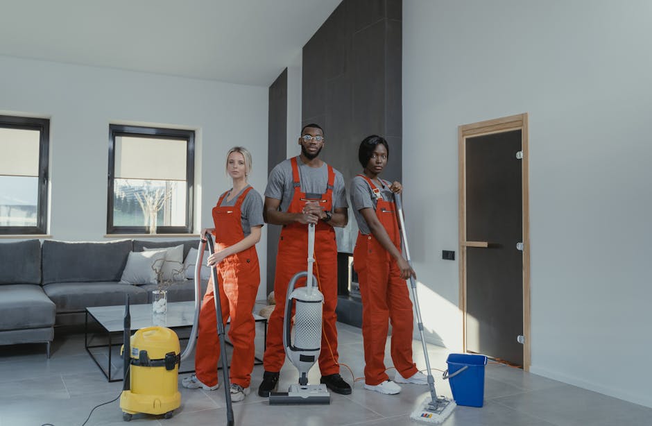 Two professional cleaners from Tottenham Green Carpet Cleaners are engaged in a domestic deep cleaning task inside a modern living room. One individual is using a handheld vacuum cleaner to remove dus