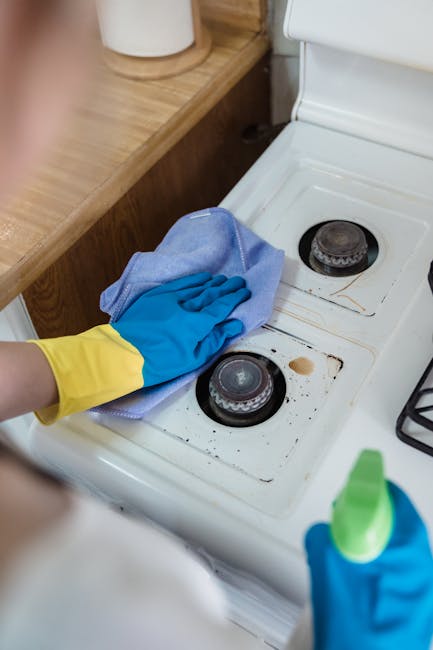 Close-up of a person's hand wearing a yellow and blue rubber glove, cleaning the stovetop of a white ceramic cooker with a blue cloth. The stovetop has two black burners, one of which shows some burnt